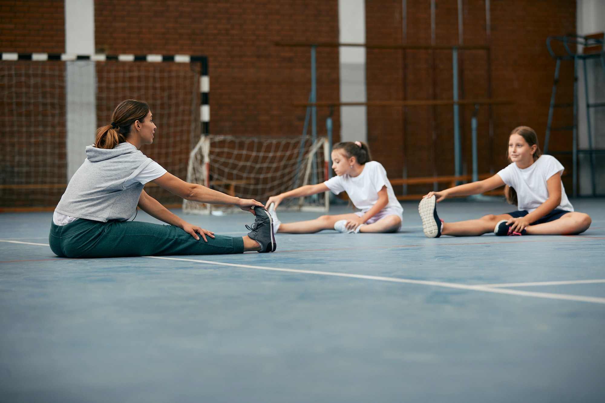 Female coach and elementary students stretching during physical education at school gym.